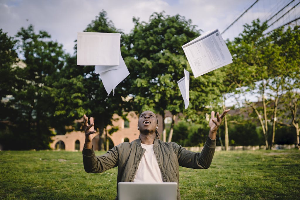 photo by ketut subiyanto Happy young black male freelancer throwing papers while celebrating successful project during remote work in green park