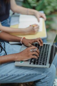 Two students studying outdoors, typing on laptops and writing notes.