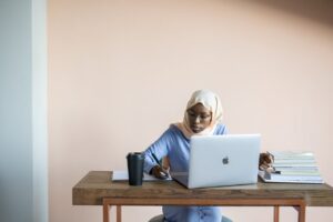 A Muslim woman in a headscarf studies diligently using a laptop at her desk with books and a cup.
