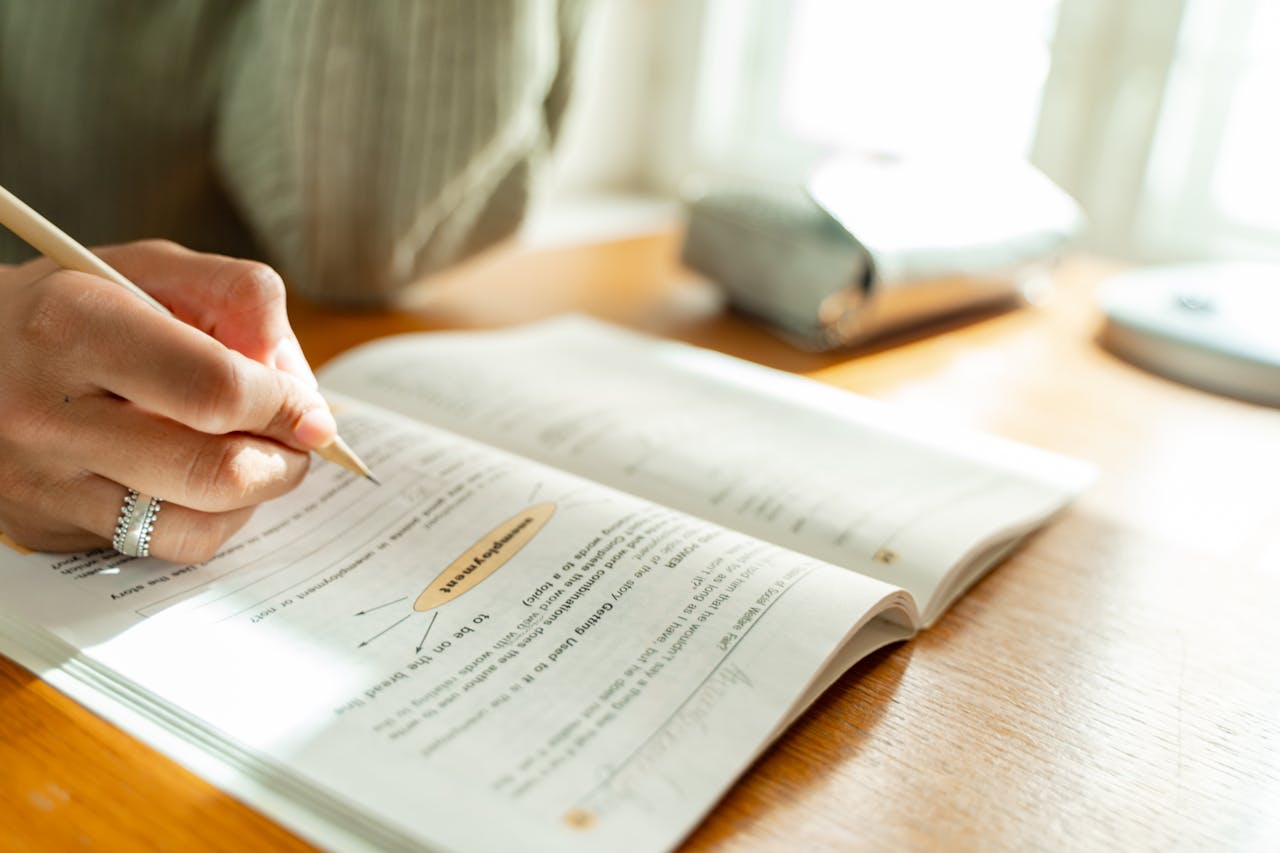 A student takes notes while studying at a sunlit desk with an open textbook.